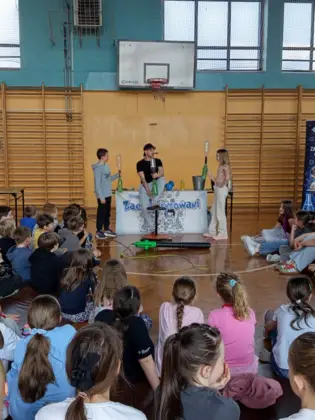 Uczniowie siedzący na sali gimnastycznej podczas pokazów fizyczno-chemicznych grupy Zaelektryzowani; na środku prowadzący i dwoje uczniów wykonujących doświadczenie.